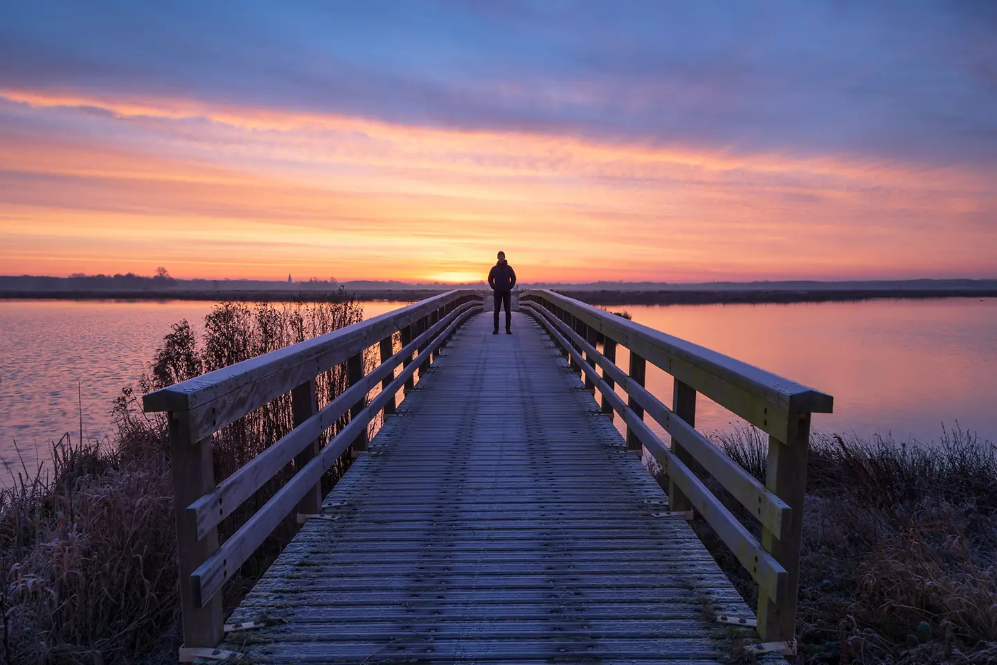 A man on a wooden bridge enjoying the view during a tranquil sunrise.
